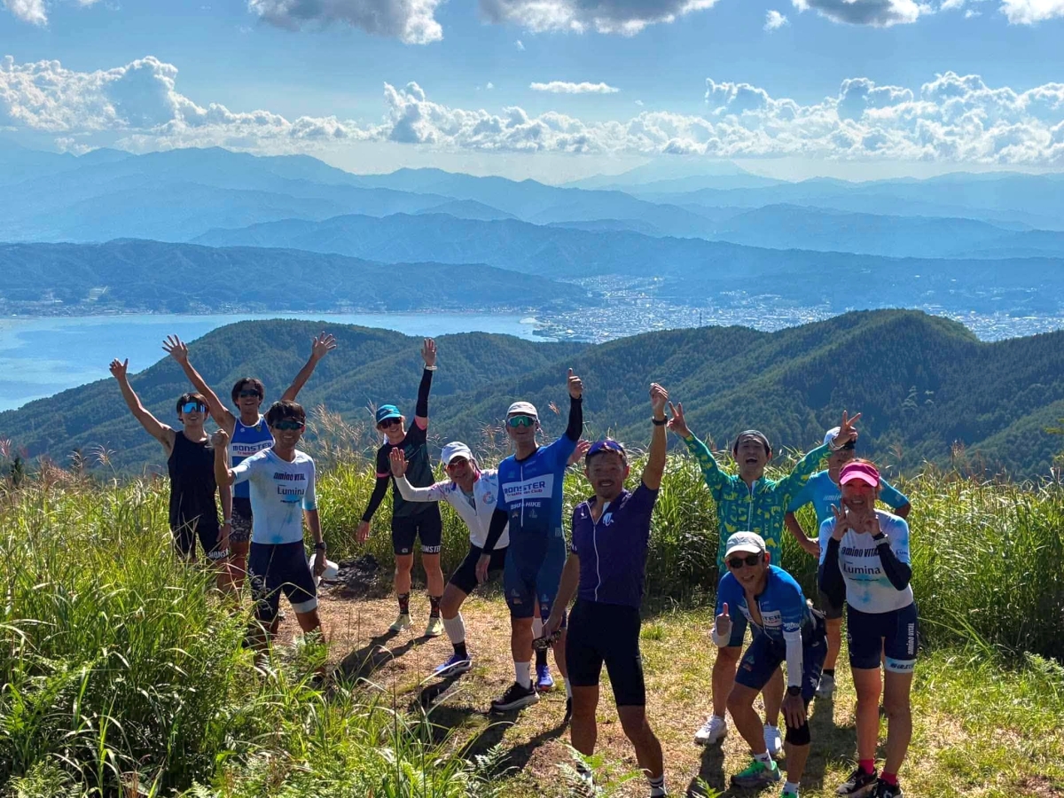 湖と山並みを見下ろす山頂で手を挙げるクラブのメンバー
